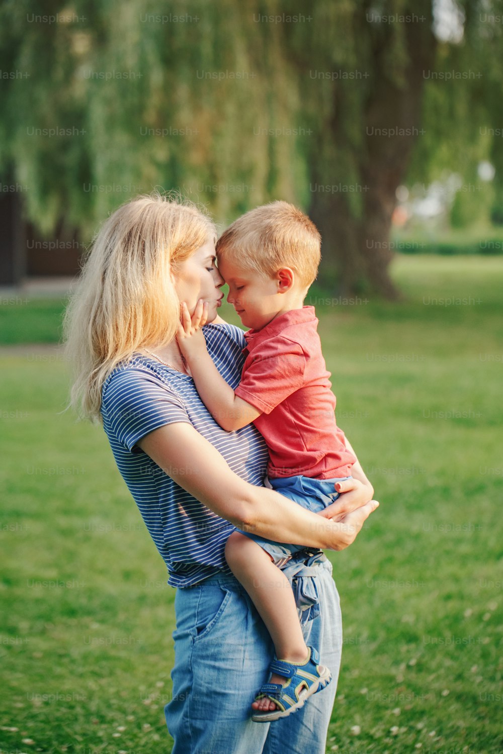 Young Caucasian mother holding boy toddler son in hands arms. Family mom kissing hugging embracing a child outdoor in park on summer day. Happy authentic family childhood lifestyle.