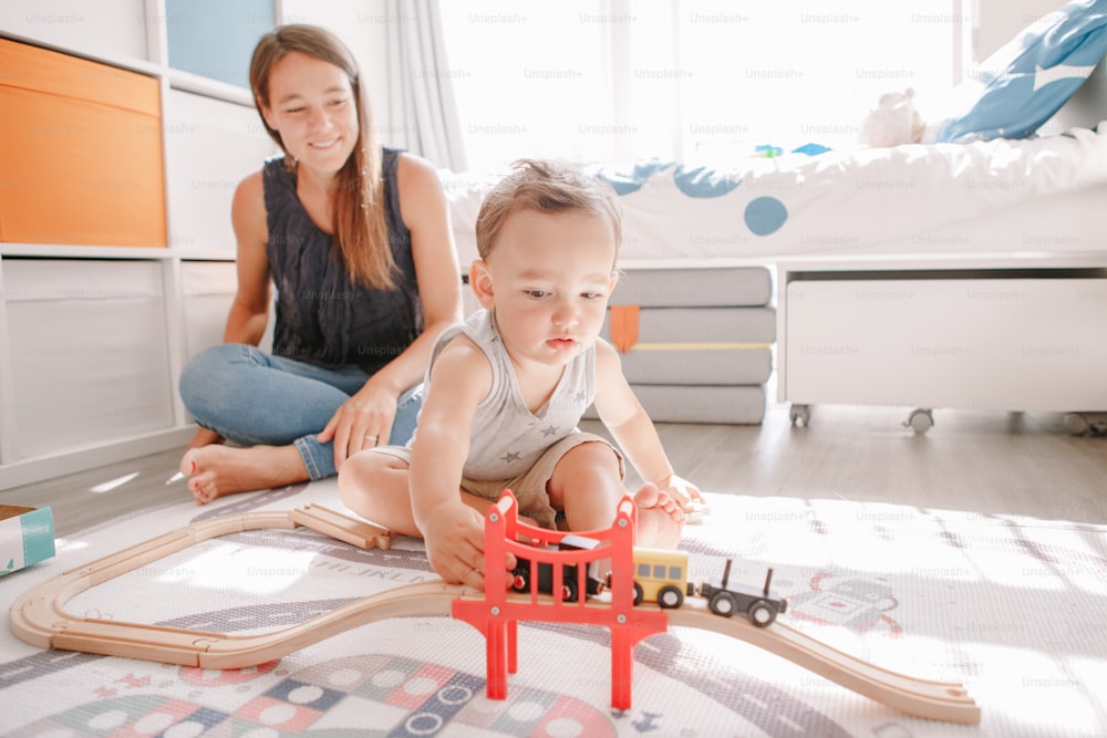 Mother and toddler boy playing with car wooden railway on floor at home. Early age education development. Kids building rail road and playing educational toy trains cars. Leisure activity for kids.
