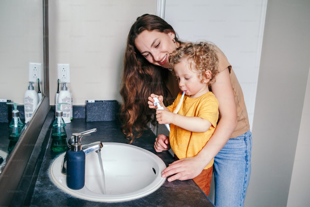 Young Caucasian mother helping boy toddler brush teeth in bathroom at home. Health hygiene and morning routine for children. Mom helping supporting teaching child to use toothbrush.