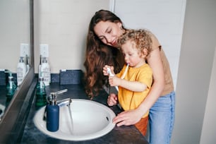 Jeune mère caucasienne aidant un garçon enfant en bas âge à se brosser les dents dans la salle de bain à la maison. Hygiène de santé et routine matinale pour les enfants. Maman aidant à soutenir l’apprentissage de l’enfant à utiliser une brosse à dents.