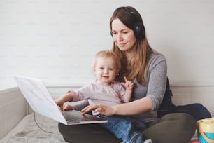 Young Caucasian mother with toddler baby working on laptop from home. Workplace of freelance woman student with kid. Online learning education or work job during self isolation, quarantine.