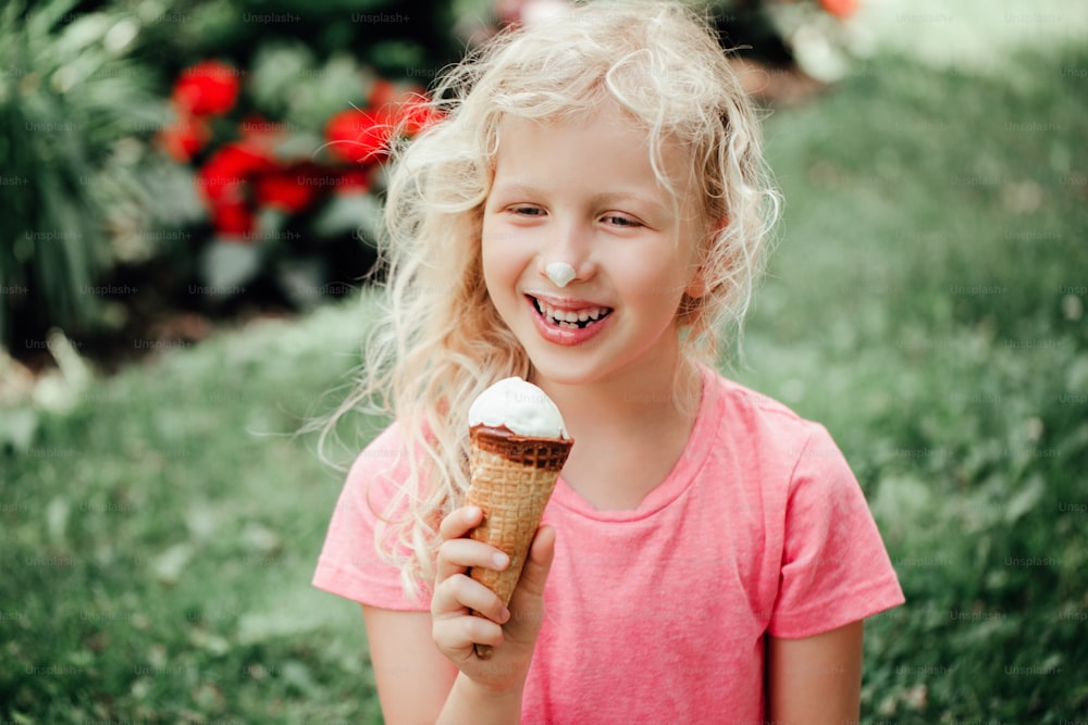 더러운 코와 우유 콧수염을 가진 귀여운 재미있는 사랑스러운 소녀가 와플 콘에서 아이스크림을 핥고 있습니다. 야외에서 맛있는 달콤하고 추운 여름 음식을 먹는 아이. 여름 냉동 식사 간식.