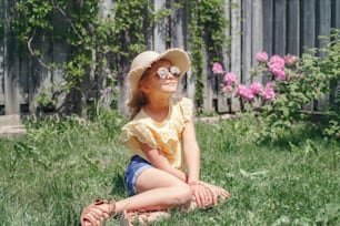 Portrait of cute adorable child girl in sunglasses and straw hat sitting on grass outdoor. Happy smiling Caucasian kid having fun at home backyard. Amazing joyful summer and lifestyle childhood.