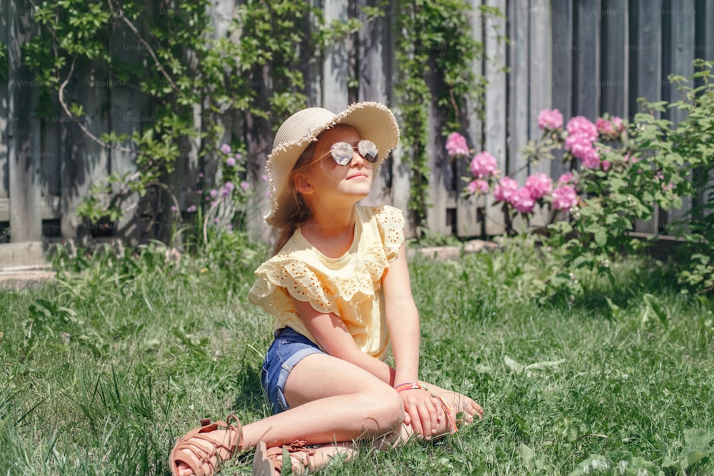 Portrait of cute adorable child girl in sunglasses and straw hat sitting on grass outdoor. Happy smiling Caucasian kid having fun at home backyard. Amazing joyful summer and lifestyle childhood.