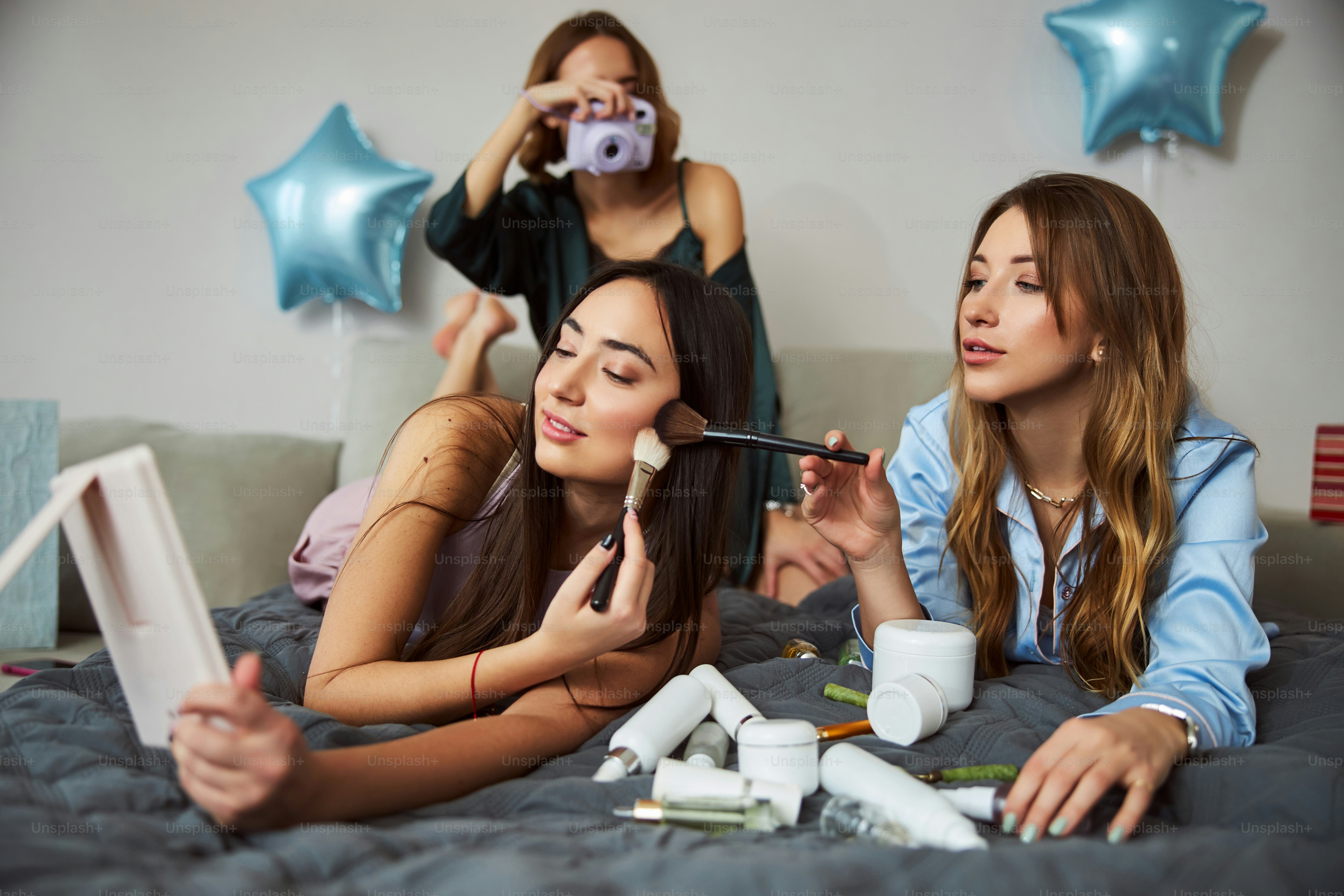 Des brune et une femme blonde photographiées par un photographe debout derrière elles pendant l’application du maquillage.