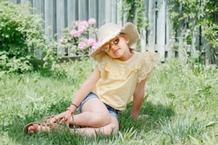 Retrato de linda niña adorable con gafas de sol y sombrero de paja sentada en la hierba al aire libre. Niño caucásico sonriente feliz divirtiéndose en el patio trasero de casa. Increíble verano alegre y estilo de vida infantil.