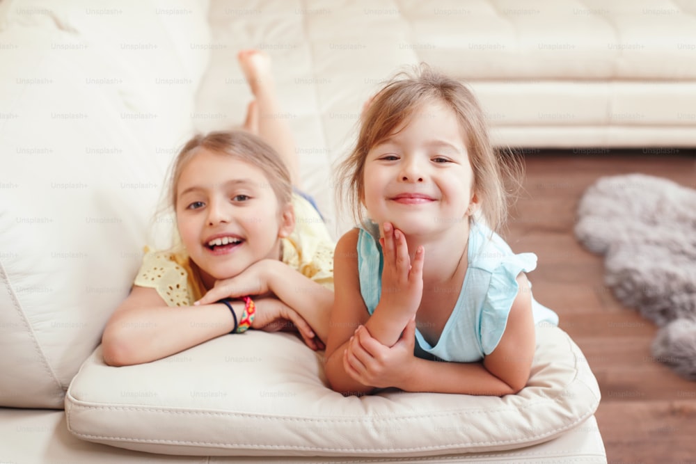 Duas meninas caucasianas fofas brincando em casa. Crianças sorridentes adoráveis deitadas no sofá juntas. Autêntico momento de vida doméstica de estilo de vida sincero. Feliz relacionamento de irmãs amigas.