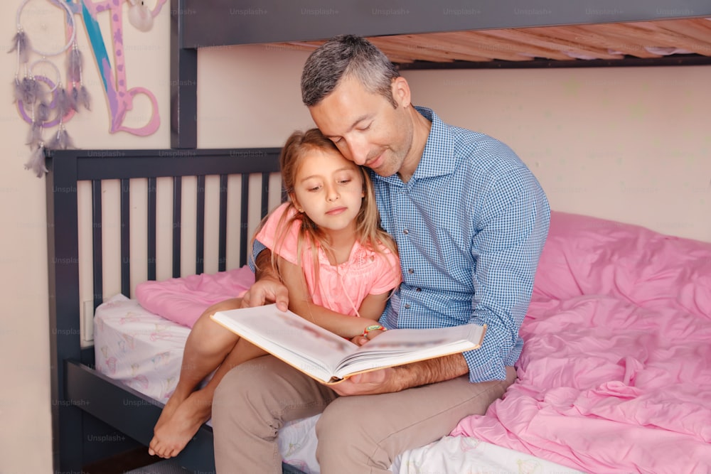 Dad reading book to pre-teen daughter girl. Happy family of two sitting on bed in bedroom. Smiling father and child at home spending time together. Education and authentic lifestyle childhood.