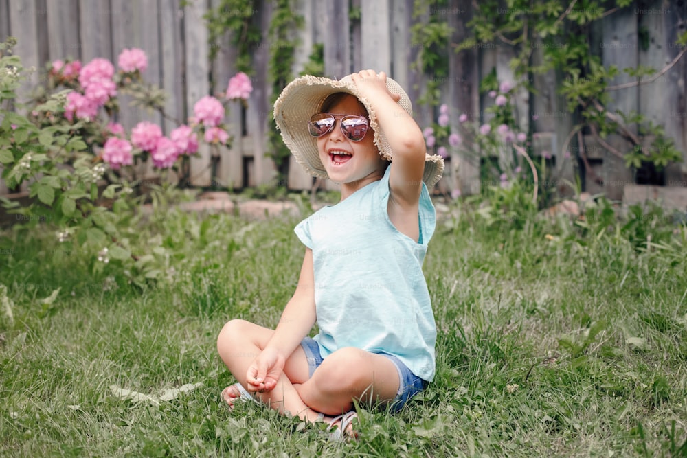 屋外の芝生の上に座っているサングラスと麦わら帽子をかぶったかわいい愛らしい子供の女の子の肖像画。自宅の裏庭で楽しんでいる幸せな笑顔の白人の子供。驚くほど楽しい夏とライフスタイルの子供時代。