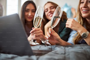 Trois jeunes femmes caucasiennes trinquent leurs verres de vin mousseux devant l’ordinateur portable