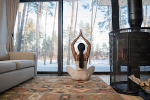 Full length mindful young woman making praying gesture while sitting in lotus position on carpet at home. Peaceful millennial girl deeply meditating, doing breathing yoga exercises alone