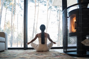 Vista posteriore della calma donna bruna multirazziale in abiti casual che medita e si siede sulla posa del loto a casa. Ragazza seduta sul pavimento di casa e guardando panorami incredibili. Concetto di yoga e benessere. Immagine