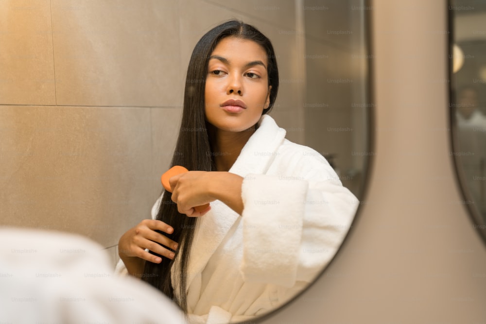 白いバスローブを着た若い自信に満ちた女性は、鏡の前に立ち、長く健康的なブルネットの髪を磨きながら、自分の反射を見ています。朝の美容ルーティンをしているミレニアル世代の女性