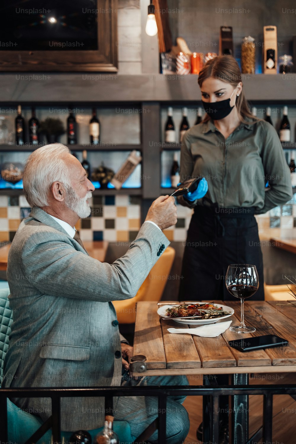 Waitress serves senior gentleman. She holds a card reader while businessman uses his credit card to pay bill. She wears a protective mask as part of security measures against the Coronavirus pandemic.
