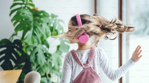 A rear view of cheerful small girl with headphones indoors at home, listening to music.