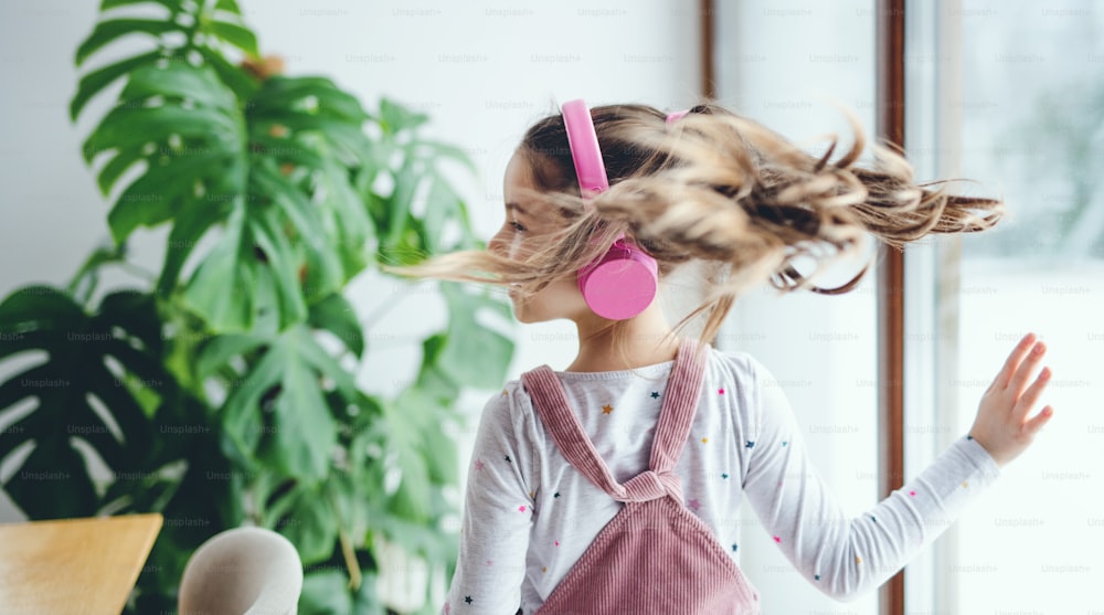 Una vista trasera de una niña pequeña y alegre con auriculares en el interior de casa, escuchando música.