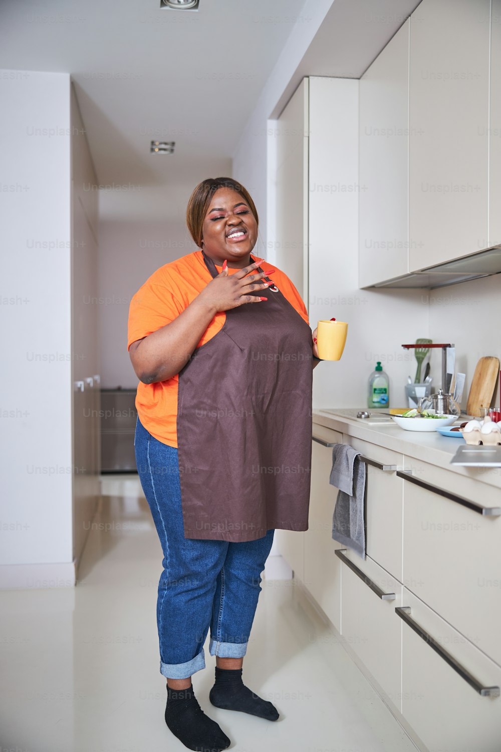 Merry Afro-American lady smiling while wearing apron and enjoying cup of coffee in her kitchen