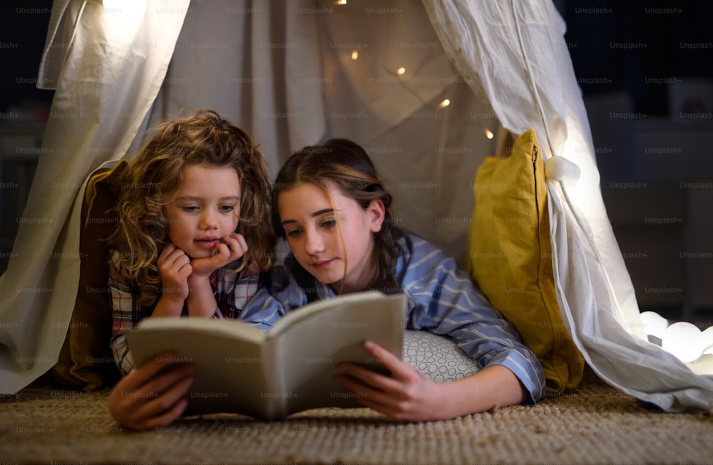 Sisters indoors at home, reading book in pajamas in tent. Lockdown concept.