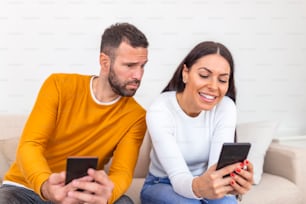 Young couple using smartphones at home. Jealous boyfriend spying his girlfriend's phone. Jealous boyfriend watching his girlfriend texting on the phone