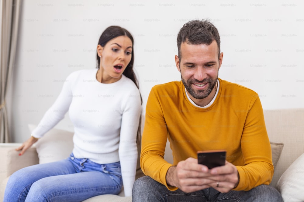 Mujer joven celosa con teléfono inteligente que mira al novio sonriente usando un teléfono inteligente en casa, concepto de problema de relación. Concepto de desconfianza