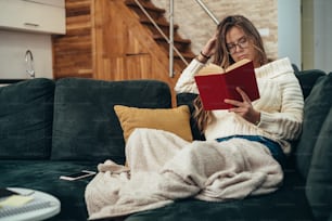 Plan d’une jeune femme lisant un livre tout en se relaxant sur le canapé à la maison
