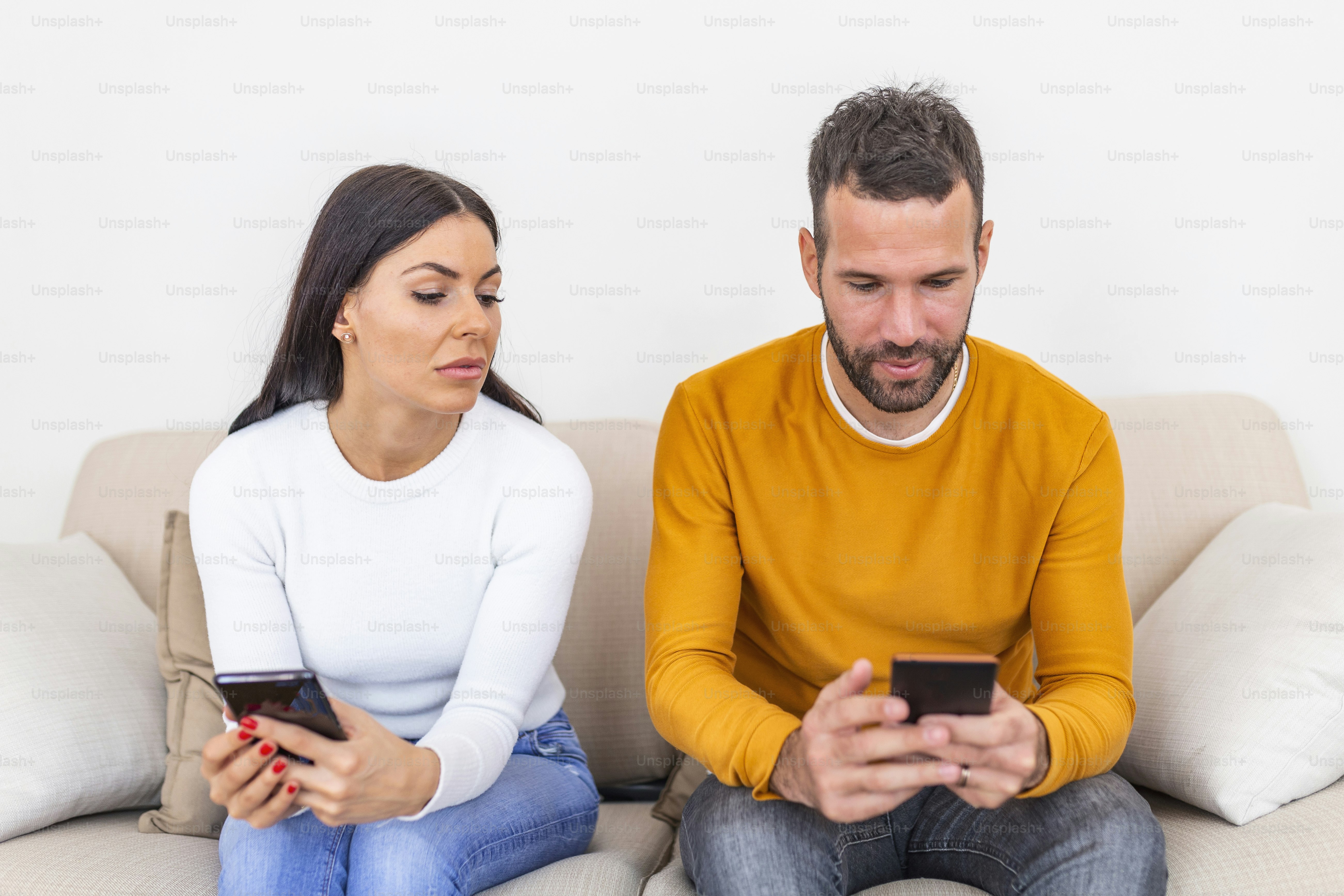 自宅でスマートフォンを使用して笑顔のボーイフレンドを見ているスマートフォンを持つ嫉妬深い若い女性、人間関係の問題のコンセプト。不信の概念