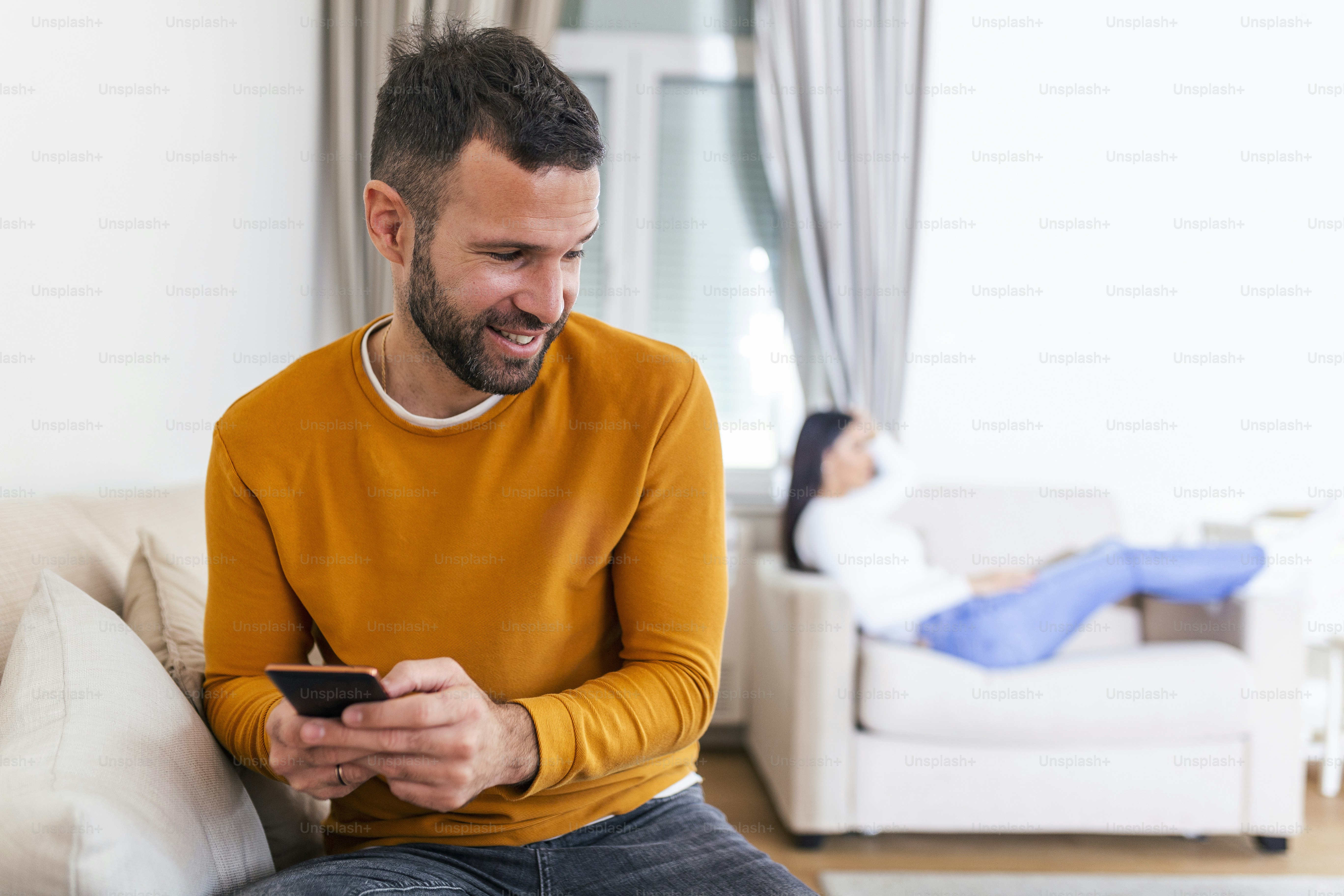 Happy smiling man turned his back to wife, reading message on phone from his lover, Woman lying on sofa in the back. Cheating and infidelity concept