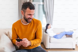 Happy smiling man turned his back to wife, reading message on phone from his lover, Woman lying on sofa in the back. Cheating and infidelity concept