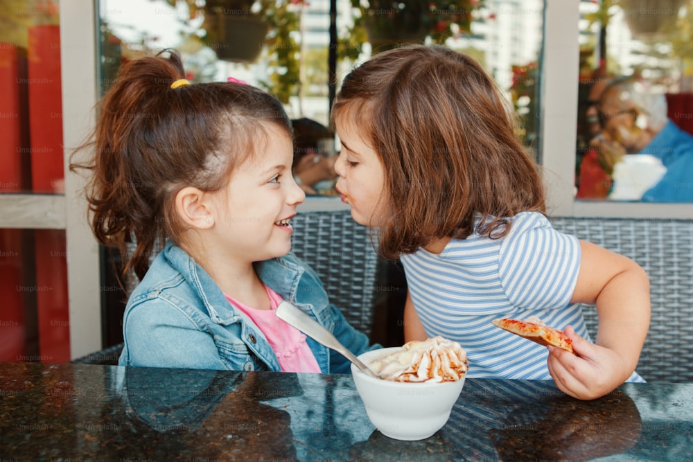 Two funny Caucasian little preschool sisters siblings kiss in cafe. Friends girls having fun together. Kids eating have breakfast in restaurant patio. Happy authentic childhood lifestyle.
