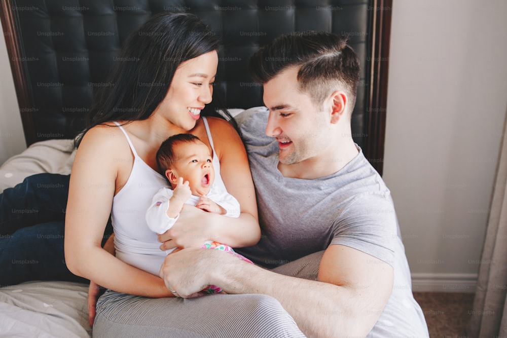 Beautiful smiling Chinese Asian mother and Caucasian father with mixed race newborn infant baby son daughter. Happy family in bedroom. Home lifestyle authentic natural moment.