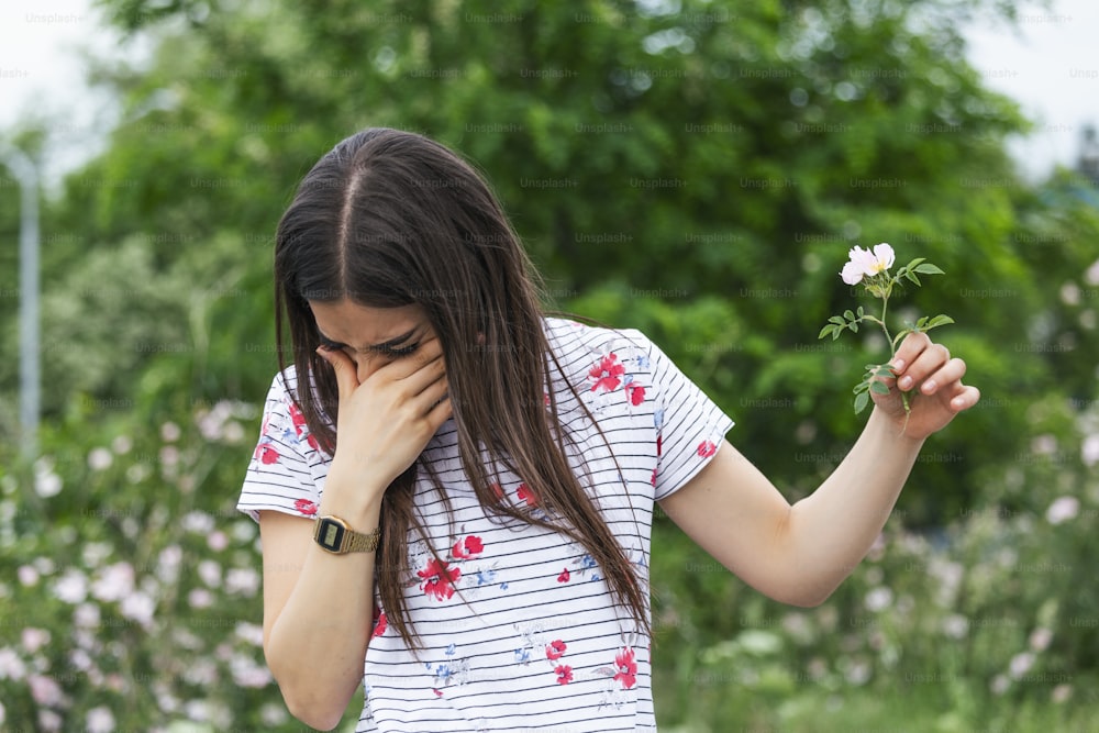 Young woman with Pollen allergy holding a flower and saying no.. Young woman with pollen and grass allergies. Flowering trees in background. Spring Seasonal allergies and health problems.