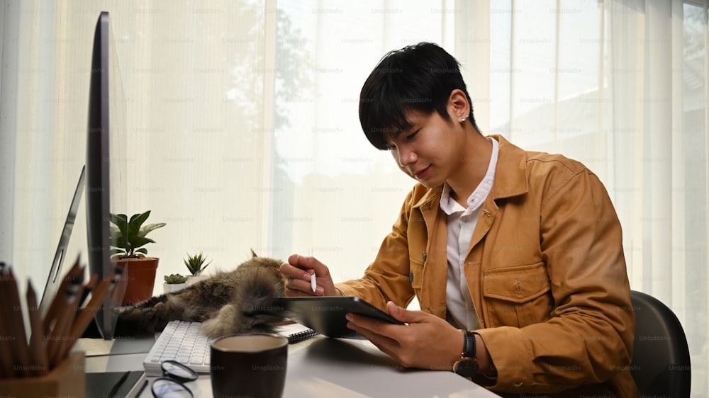 Diseñador joven sonriente usando tableta digital mientras está sentado en una cómoda oficina en casa.