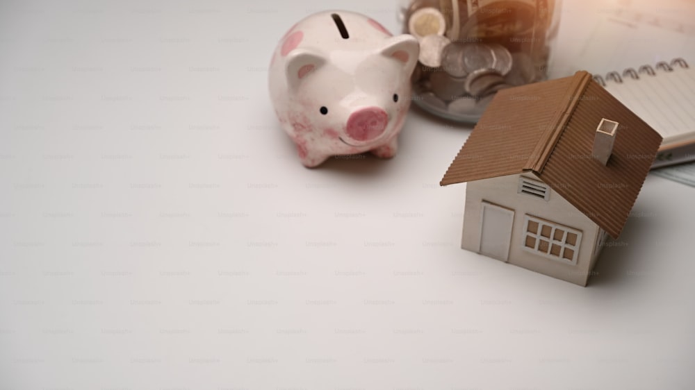 돼지 저금통, 동전 및 흰색 테이블에 집 모델. 미래를 위해 돈을 저축하고, 집이나 부동산 시장을 사기 위해 저축하십시오.