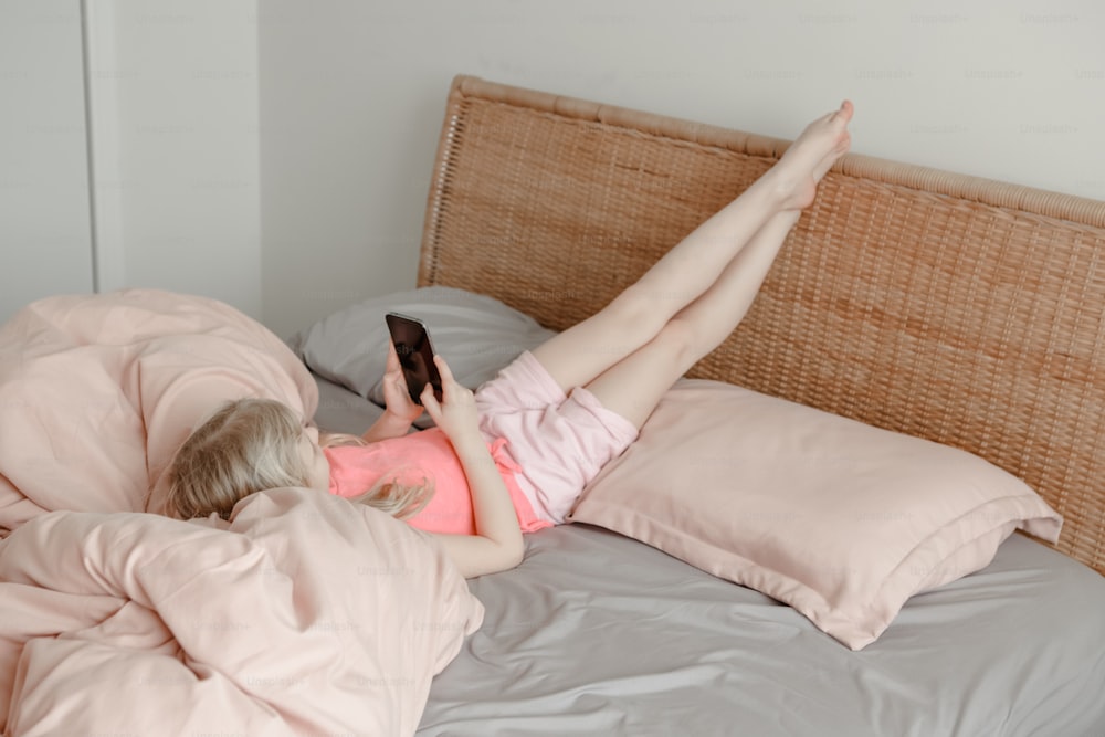 Fille allongée dans son lit à la maison et discutant avec des amis sur un smartphone en ligne. Enfant enfant parlant en tapant au téléphone dans les médias sociaux. Auto-isolement sur la quarantaine coronavirus covid-19. Dépendance au temps d’écran.