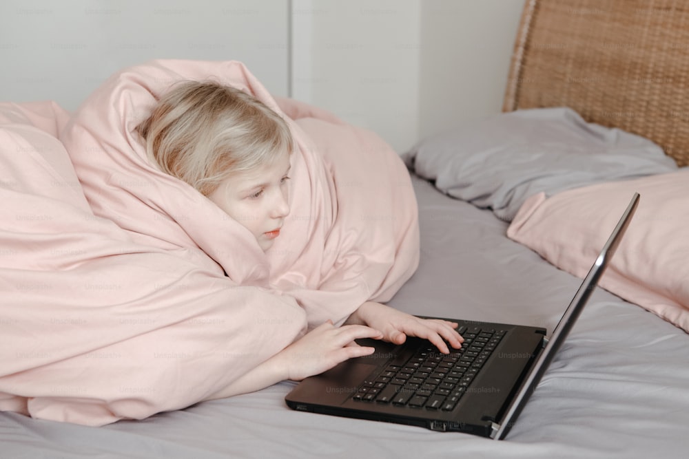Bored girl lying in bed under blanket and learning in virtual online school class. Kid working on laptop Internet at home. Child using wireless technology education. Homeschooling for children.