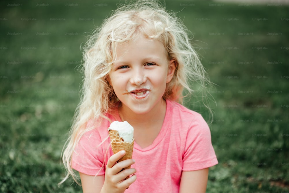 汚れた鼻とミルクの口ひげを生やしたかわいくて面白い愛らしい女の子が、ワッフルコーンからアイスクリームを舐めて食べています。屋外で美味しくて冷たい夏の食べ物を食べる子供。夏の冷凍食品スナック。