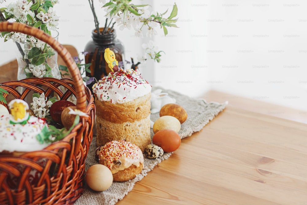 Homemade Easter bread, modern natural dyed eggs, wicker basket, bunny and blooming spring flowers on rustic table. Happy Easter. Traditional easter food for church blessing and festive dinner