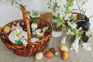 Stylish easter eggs, homemade Easter bread, delicious traditional Easter food in wicker basket and blooming spring flowers on linen napkin on rustic table. Happy Easter! Festive breakfast