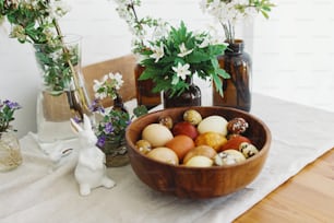 Stylish easter eggs in wooden bowl on rustic table with bunny figurines and spring blooming flowers. Natural dyed eggs in yellow and red colors. Happy Easter!