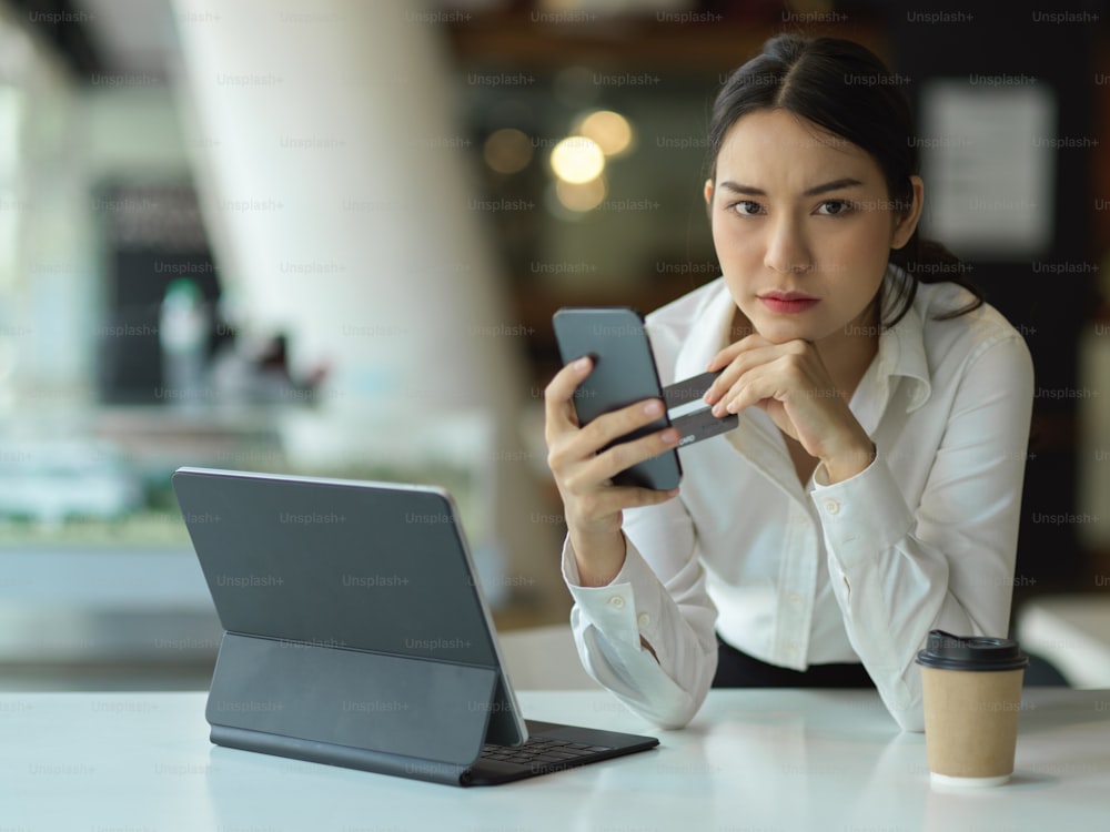 Portrait of female office worker feeling stressed and looking to camera while online paying with smartphone and credit card