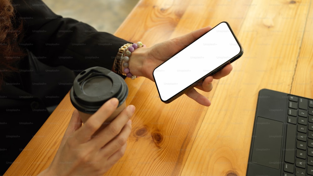 Cropped shot of businesswoman hand holding smartphone and paper cup while relaxing at workplace, clipping path
