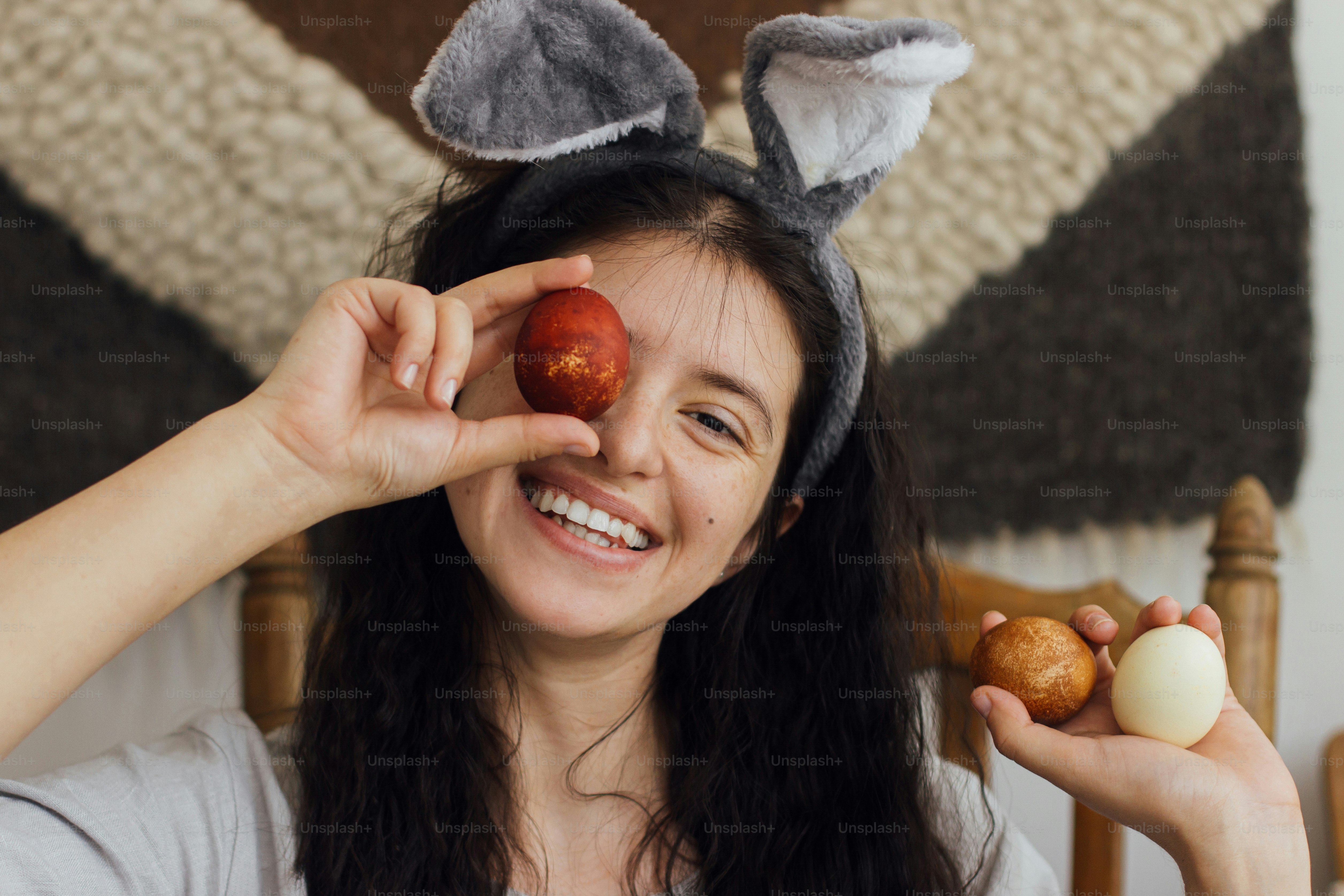 Happy young woman in bunny ears and linen dress holding easter eggs at eyes and smiling on background of rustic room. Happy Easter! Natural dyed easter eggs in hands, funny moments