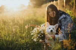 Cute white puppy with daisy flowers in warm sunset light in summer meadow. Stylish young woman hugging adorable fluffy puppy with  bouquet of wildflowers. Summertime. Adoption concept