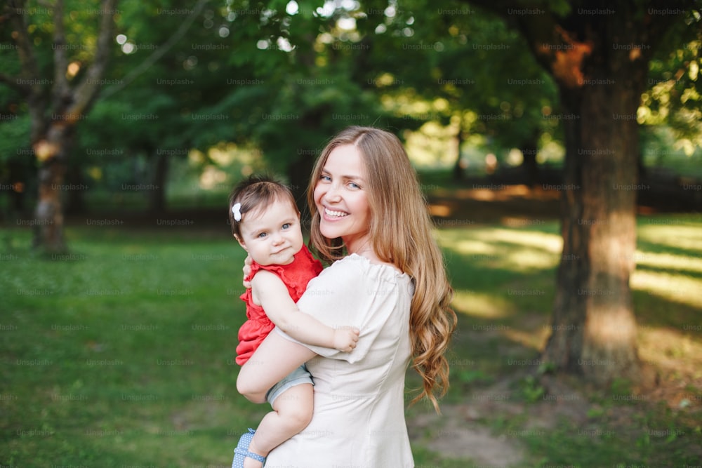 Fête des mères. Jeune mère caucasienne souriante et fille en bas âge fille s’étreignant dans le parc. Maman embrassant un bébé enfant un jour d’été à l’extérieur. Heureux et authentique style de vie d’enfance en famille.