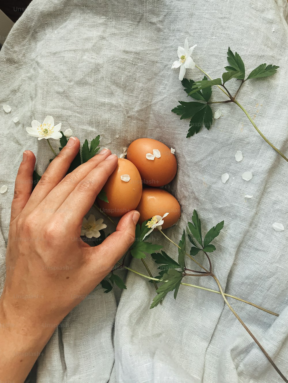 Hand holding natural easter eggs and spring flowers on background of rustic linen dress. Happy Easter! Woman holding natural eggs. Aesthetic holiday, top view