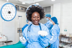 African American Plastic surgeon woman holding silicon breast implants in surgery room interior. Cosmetic surgery concept