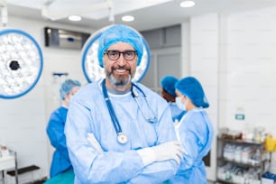 Retrato de un cirujano masculino de pie con los brazos cruzados en el quirófano del hospital. Los cirujanos del equipo están realizando una operación, el médico de mediana edad está mirando a la cámara, en un quirófano moderno