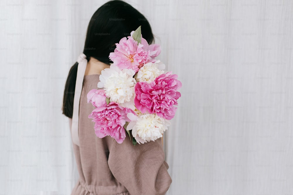 Beautiful stylish woman in linen dress holding peony bouquet on background of white fabric. Slow life. Young female in boho rustic dress hiding face with pink and white peonies. Aesthetic moment