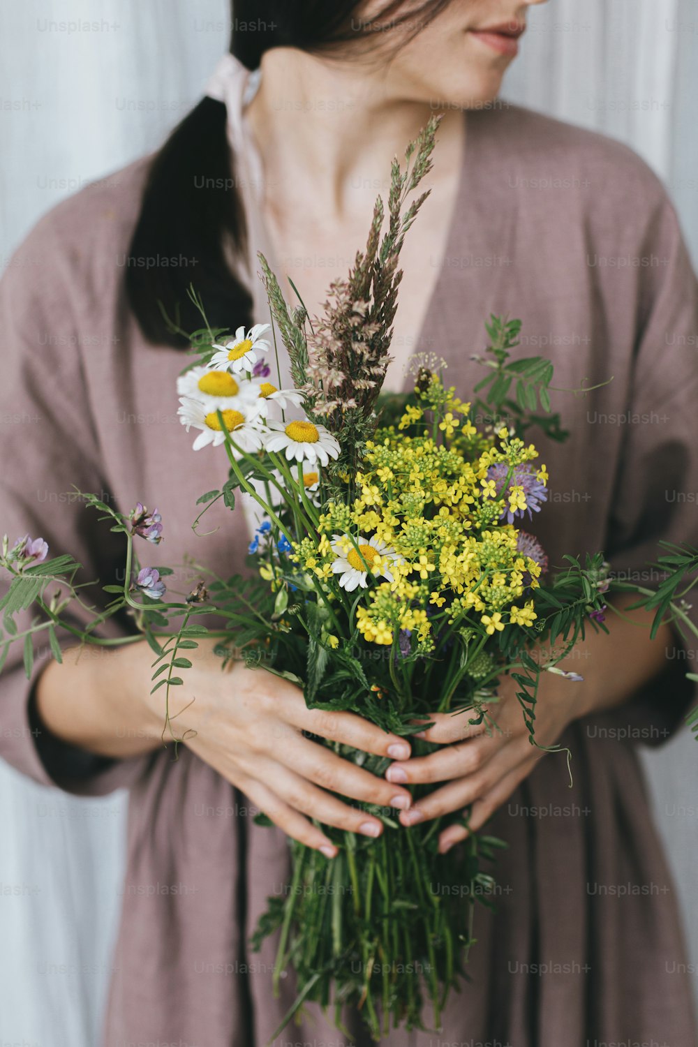 Stylish woman in linen dress holding wildflowers bouquet on background of pastel fabric. Simple slow living. Young female in boho rustic dress with white and yellow meadow flowers in hands