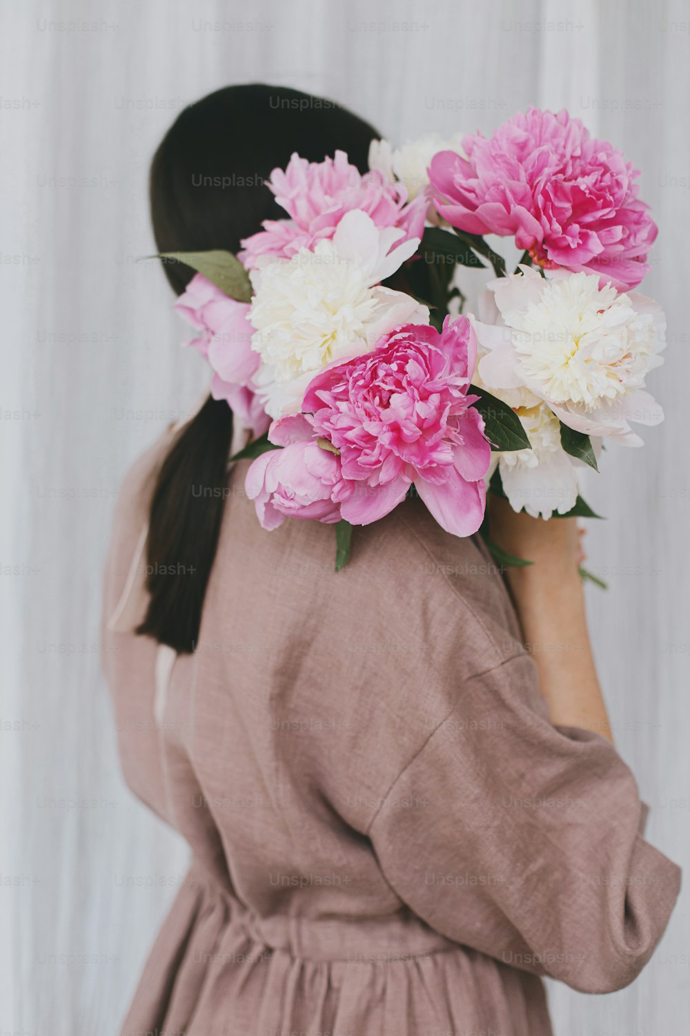 Stylish woman in linen dress holding peony bouquet on background of white fabric. Slow life, little happy things. Young female in boho rustic dress hiding face with pink and white peonies.